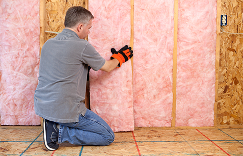 Technician kneeling on unfinished floor, installing pink fiberglass batt insulation in an unfinished wall.