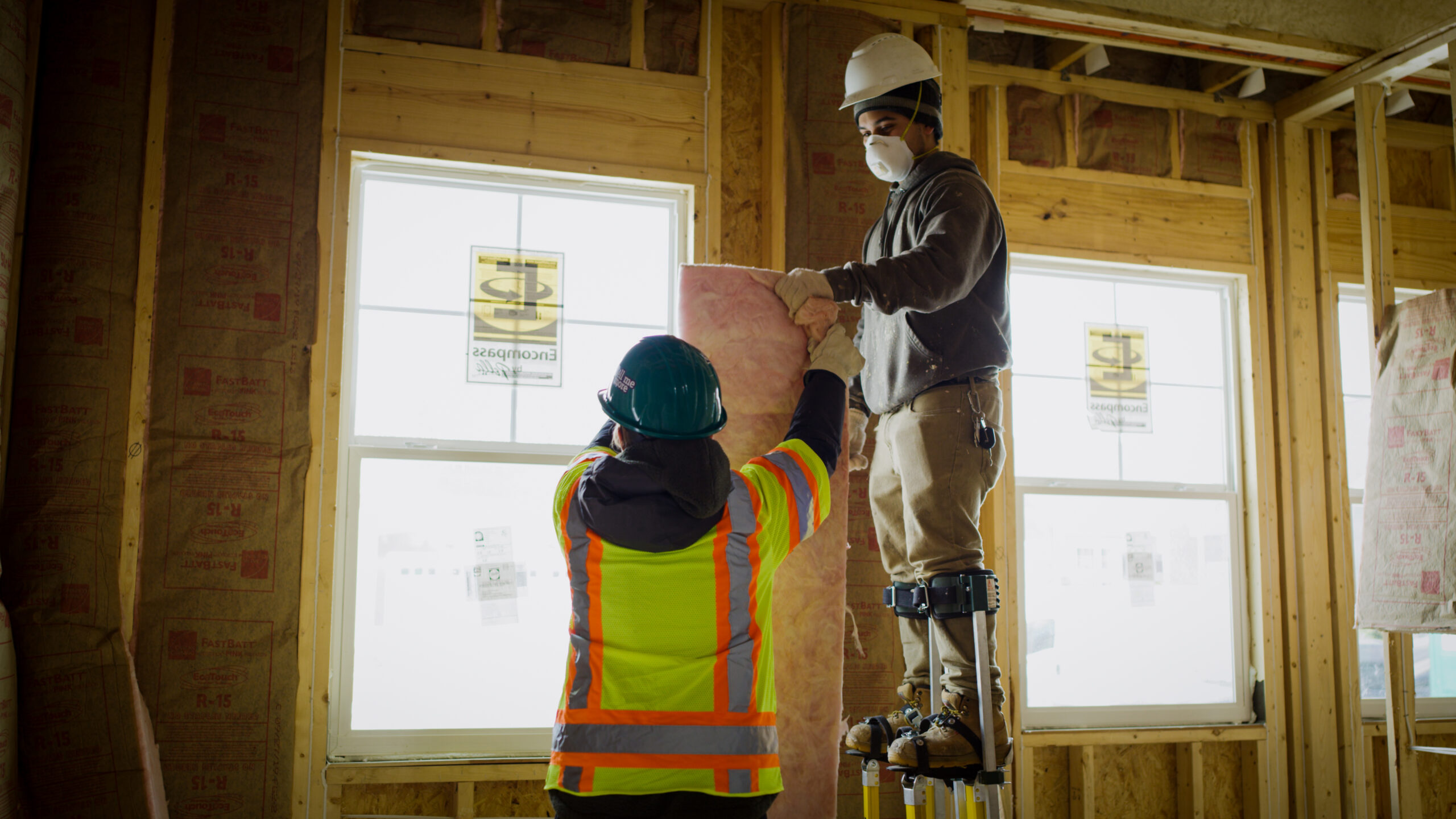 Two contractors installing pink fiberglass batt insulation in new construction.