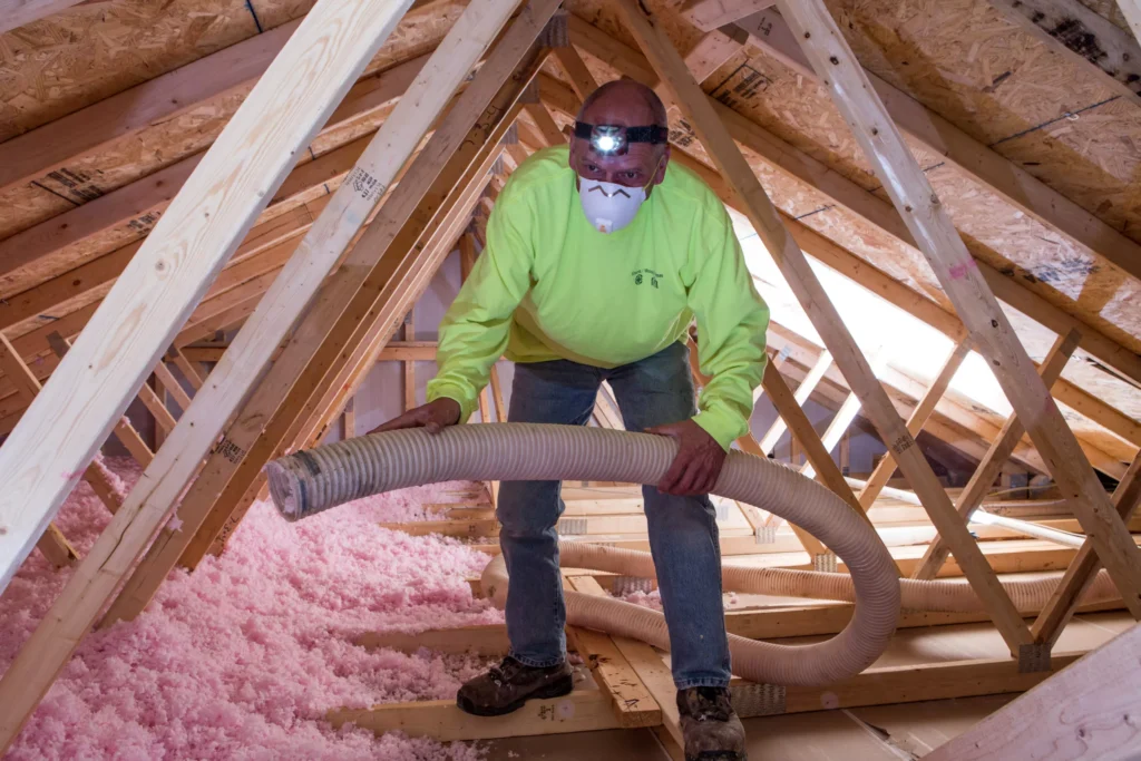 Technician installing loose-fill pink fiberglass insulation in an unfinished attic.